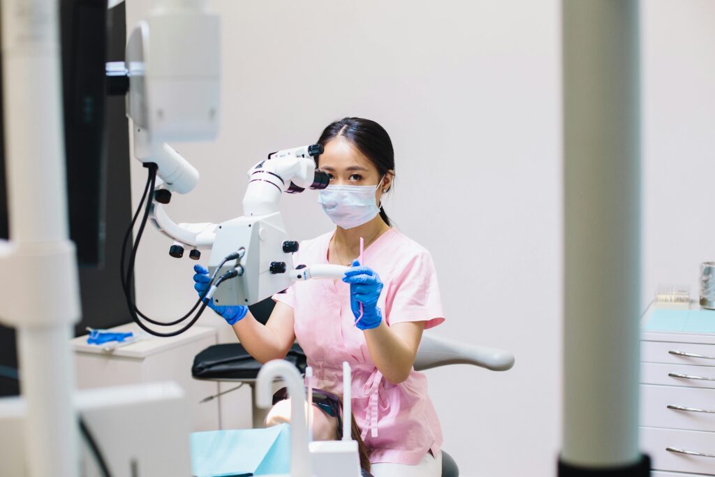 A female dentist wearing a mask uses a microscope in a dental clinic setting for precision treatment.
