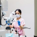 A female dentist wearing a mask uses a microscope in a dental clinic setting for precision treatment.