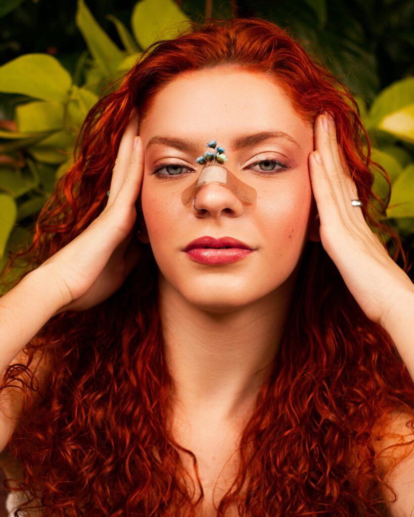 Artistic portrait of a young woman with a flower on a band-aid placed on her nose, surrounded by lush greenery.